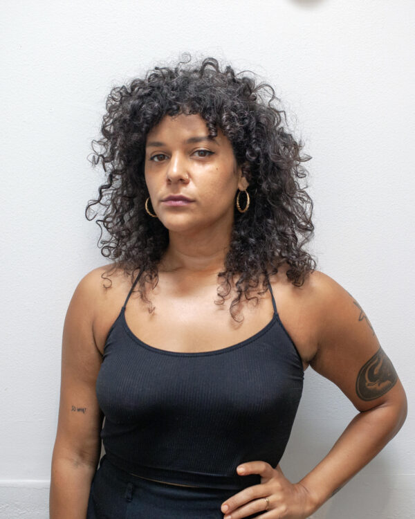 An AfroIndigenous person with curly hair and a black tanktop and skirt stares at the camera