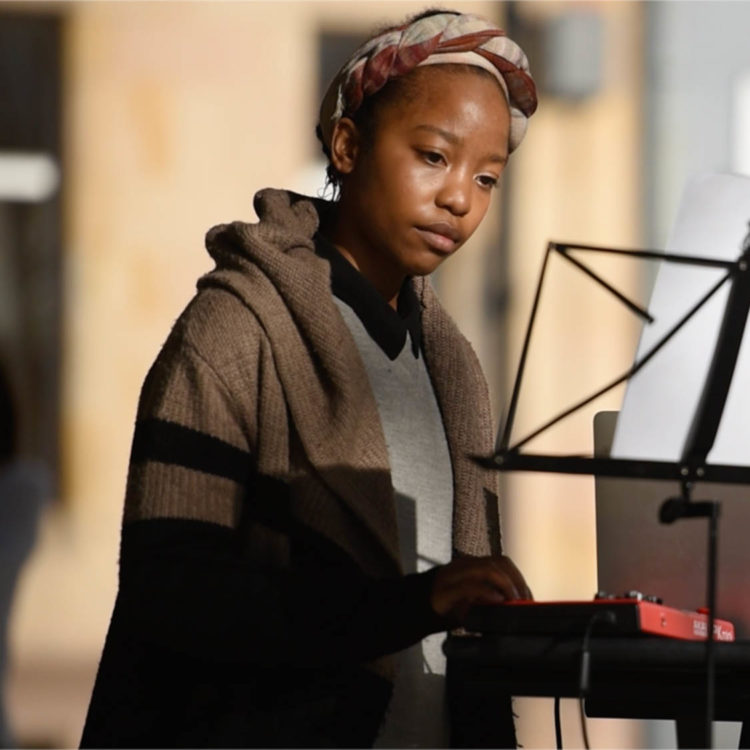 A woman with a head scarf faces a music stand, focused and attentive as she touches a keyboard.