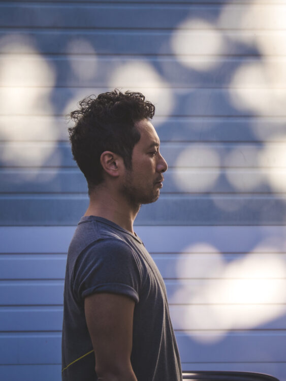 Profile image of artist Takahiro Yamamoto in front of an outdoor garage door in a blue light with scattered warm lights bouncing off.