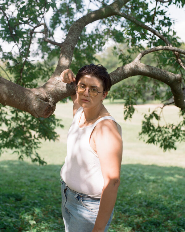 A latinx non-binary individual with short black hair wearing a white tank top and jeans stands outside in speckled sunlight in front of a tree at an empty park. Their right arm and hand rest loosely on a tree branch behind them while they look straight into the camera.
