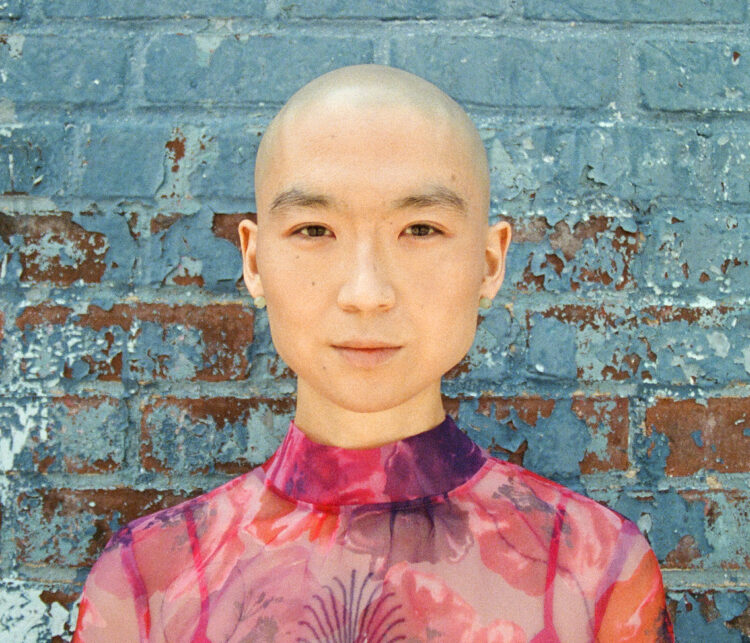 yuniya edi kwon looks into the camera in front of a bright painted blue brick wall. She has a shaved head and is wearing jade pearl earrings and a pink-red semi-sheer dress with a floral pattern.