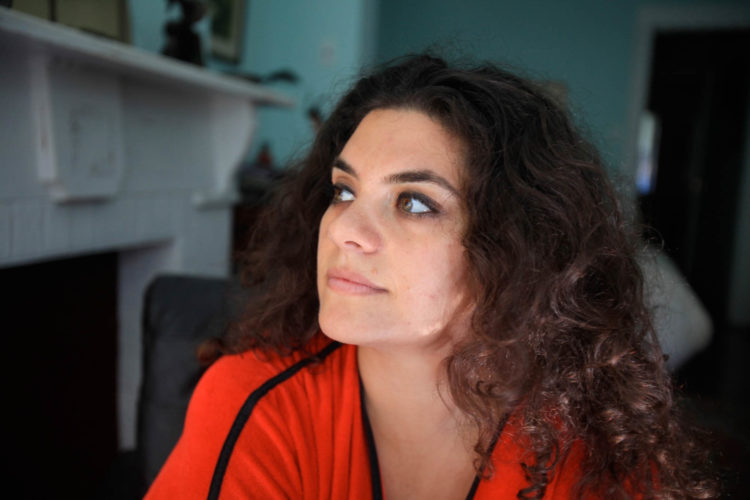 A brown haired woman, wearing a red top sits looking to her right with a light turquoise room in the background.