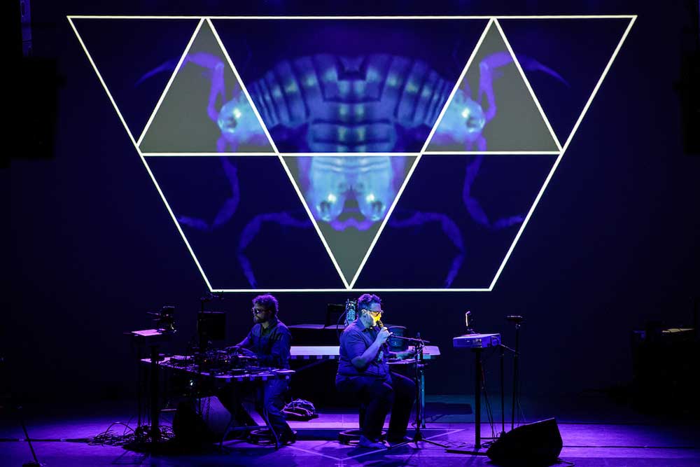 Two figures sit behind synthesizers in front of a quadrangle display above them on a stage. The stage is lit by purple and blue light.
