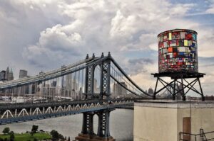 The Manhattan Bridge pictured in front of clouds and the New York City skyline. A multi-color water-tower stands in the foreground.