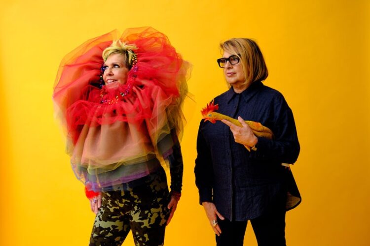 Two woman stand against a yellow background. One wears a tutu over her neck and the other holds a rubber chicken.