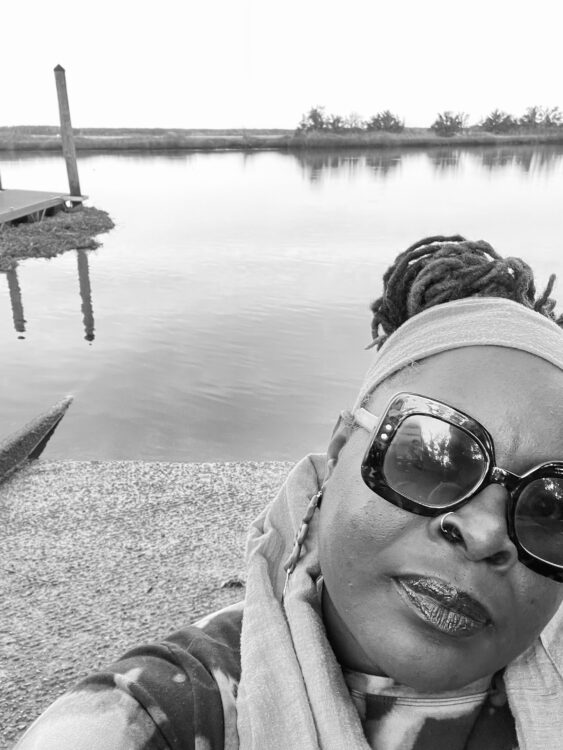 A black and white selfie of artist Ebony Golden pictured in front of a body of water. 