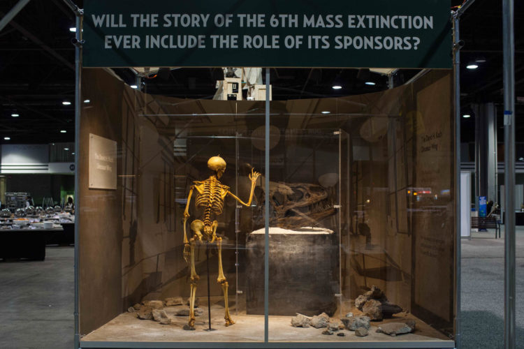 A diorama in a convention center at the American Alliance of Museum’s annual conference depicts a skeleton contemplating a display case of an allosaurus skull in a room with a plaque that says David H. Koch Dinosaur Wing. A caption overhead reads Will the Story of the 6th Mass Extinction Ever Include the Role of Its Biggest Sponsors?