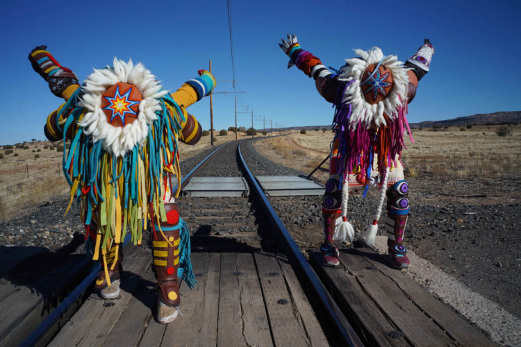Two figures wearing full regalia face the camera with full face covering regalia, arms are raised behind them as if to jump. They are exterior on train tracks.