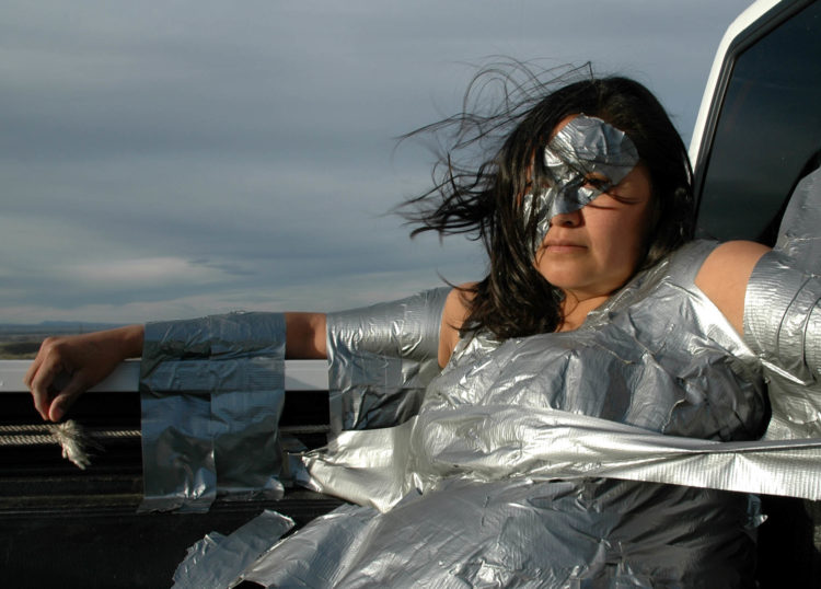 A woman with dark hair is reclined back in the bed of a pickup truck covered in duct tape with a blue and grey cloudy sky in the background.